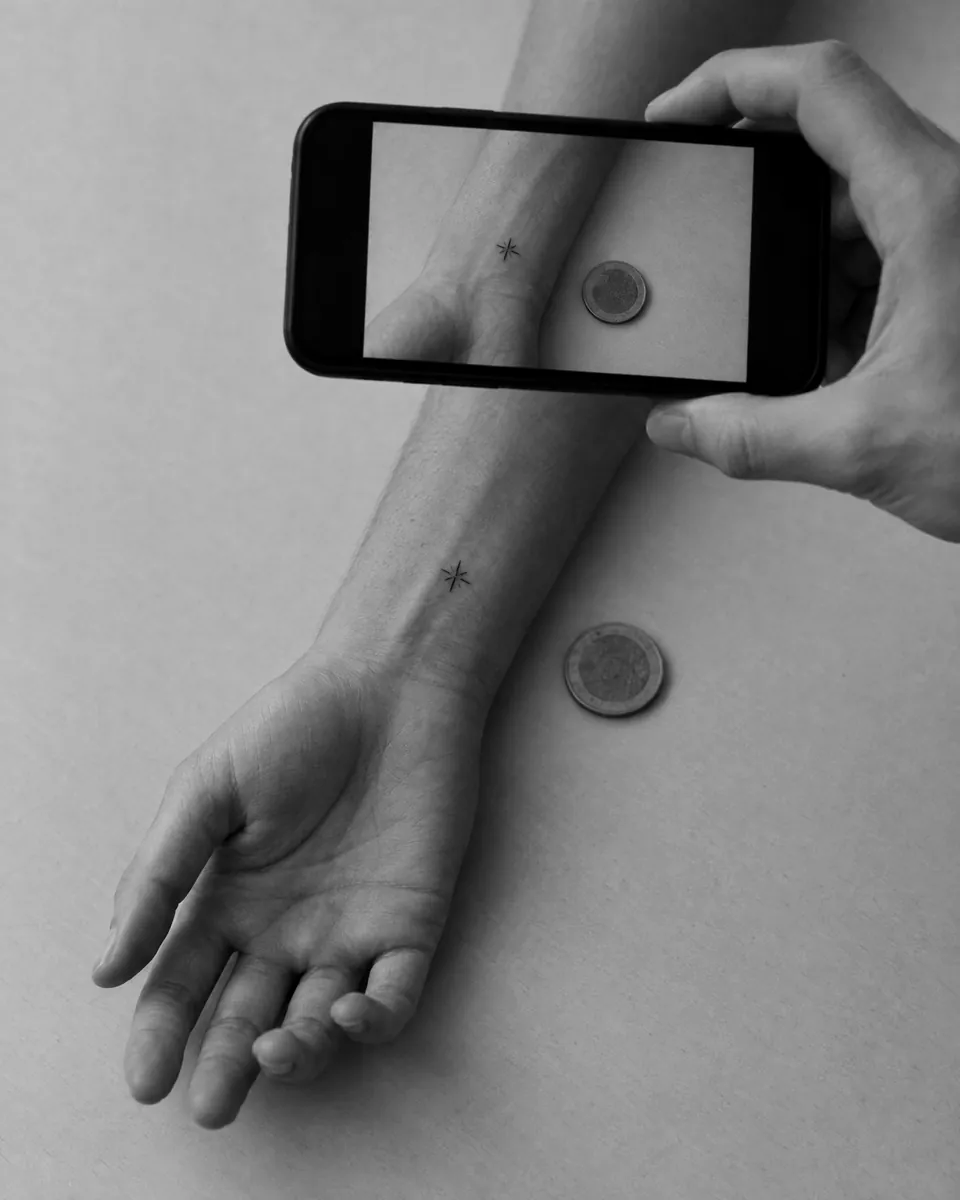 Una mano fotografía con el móvil un tatuaje pequeño en la muñeca con una moneda al lado